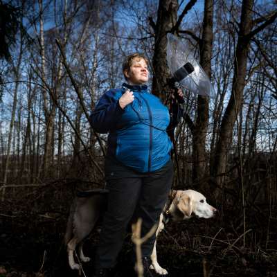 Nainen sinisessä takissa seisoo metsässä. Kädessään hänellä on parabolimikrofoni. Hänen takanaan seisoo vaalea labradorinnoutaja.