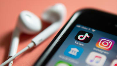 En telefon och ett par hörlurar ligger på ett persikofärgat bord. På telefonen syns olika appar, bland annat Tiktok och Instagram.