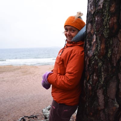 Ilse Klockars iklädd orange jacka tittar in i kameran och ler. Hon lutar mot en tall vid sandstranden i Hangö.
