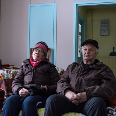 Tummanruskeisiin talvitakkeihin pukeutuneet Ritva ja Antero Kyllönen istuvat sohvalla kädet sylissä kesämökillään Kuhmon Vartiuksessa.