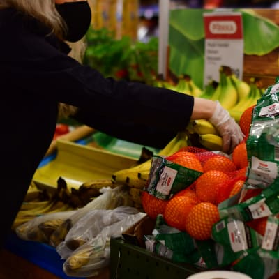 Ett butiksbiträde ordnar bananer och apelsiner med plasthandskar på.