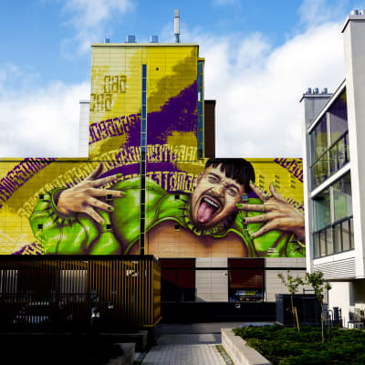Käärijä i grön bolero visar tungan på en stor mural i Dickursby, Vanda. 