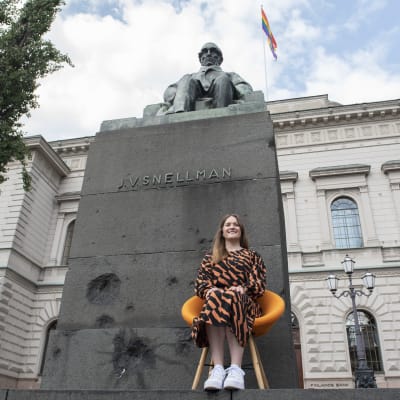 Danske Bankin pääanalyytikko Minna Kuusisto istuu oranssilla tuolilla Suomen Pankin edustalla olevan J.V.Snellmannin patsaan edessä.