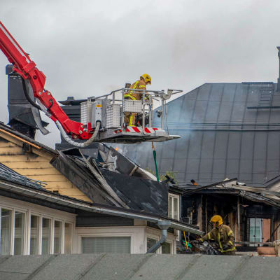 Räddningspersonal släcker brand med lyftkran. I bilden syns ett upprivet förkolnat hustak. 