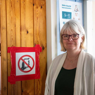 En vithårig kvinna står bredvid en skylt om telefonförbud.
