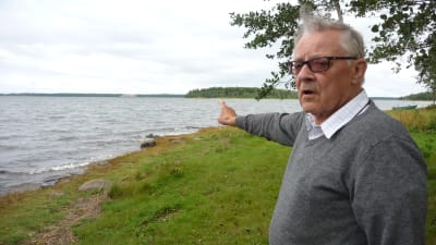 Håkan Södergård står vid stranden och pekar ut mot Storgrynnan i Pjelax som har förstörts av skarvar.