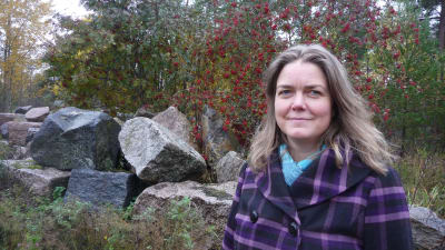 En kvinna med lila jacka står framför stora stenar och en rönn med röda bär.