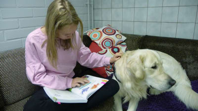 En flicka sitter med en hund i en soffa och läser bok.