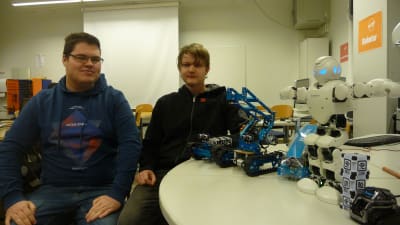Två manliga studerande sitter bredvid ett bord med robotar uppradade på det.
