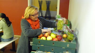 En blond kvinna står och plockar upp frukt ur en låda.