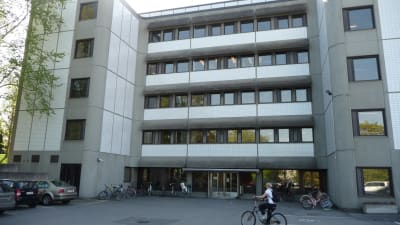 En studerande i vit t-skjorta cyklar fram till studenthälsovårdens hus i Åbo är ett grått höghus i fem våningar.