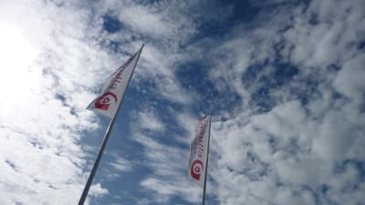 Två avlånga vita flaggor med röd text: Kårkulla, vajar i en flaggstång en solig dag med moln.