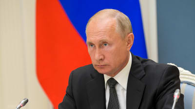 Vladimir Putin sitter vid ett bord med mikrofon framför sig och med en allvarlig min.