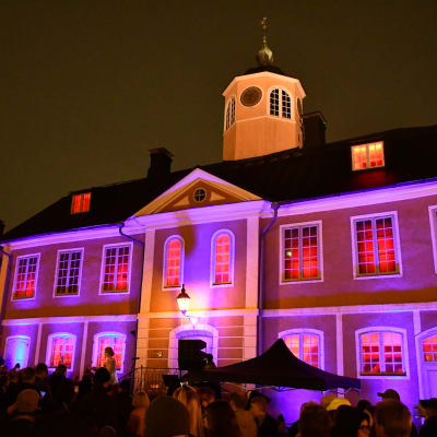 En gammal rådhusbyggnad är upplyst med röda lampor. Framför står en stor folkmassa.