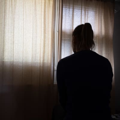 Nuori nainen istuu pimeässä huoneessa missä verhot ovat kiinni.