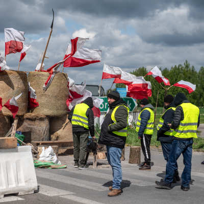 Några män i reflexväst står med händerna i fickorna framför fem höbalar med tiotals polska flaggor instuckna i sig.