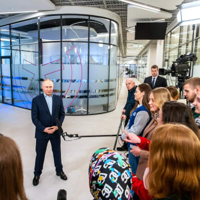Vladimir Putin seisoo keskellä lattiaa tummassa puvussa ilman kravattia kädet ristissä mahan päällä. Etualalla on joukko opiskelijoita. Taustalla seisoo ryhmä virkamiehiä, muun muassa Moskovan pormestari Sergei Sobjanin.