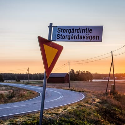 En skylt med texten Storgårdsvägen, i bakgrunden syns åkrar och en väg i soluppgången