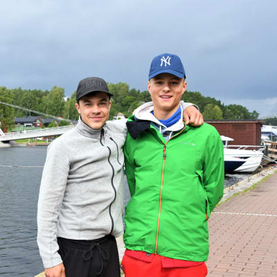 Personen till vänster har på sig en grå tröja och en svart keps och håller armen runt personen till höger. Personen till höger har på sig röda byxor, en grön jacka och blå keps. I bakgrunden ser man en bro. 