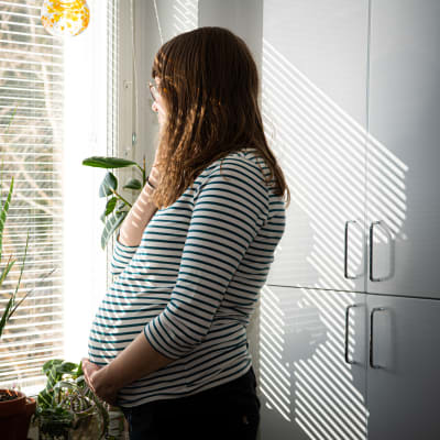 En gravid kvinna står med handen under sin stora mage och tittar ut genom ett fönster. 