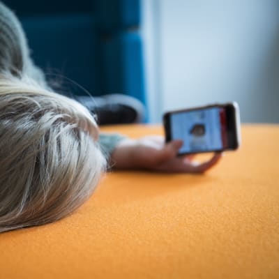 Tyttö makaa sohvalla ja katsoo kännykän näyttöä