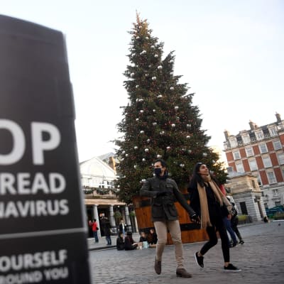 Bild på skylt där det står stoppa spridningen av coronaviruset, och i bakgrunden ett par som går hand i hand vid en stor julgran på julmarknad i London.