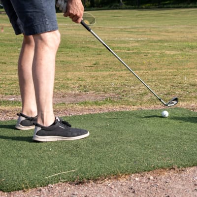 Någon håller på att slå en golfboll med golfklubba. I bilden syns tvp ben, golfboll, golfklubba och golfbanan i bnakgrunden. 