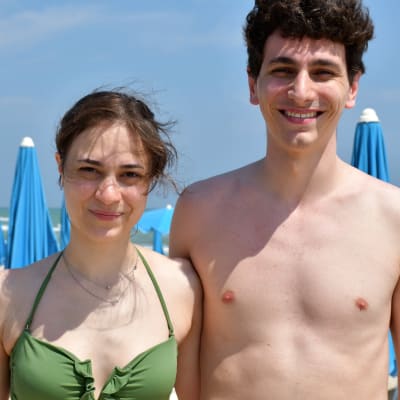 En ung kvinna och hennes kille står och håller om varandra på stranden och tittar in i kameran. Hon har en grön bikini på sig, han är barbröstad. I bakgrunden syns solstolar.