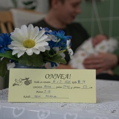 En gul lapp med texten "onnea" på och information om den nyfödda. Pappan och sonen syns i bakgrunden då de sitter i gungstolen. 