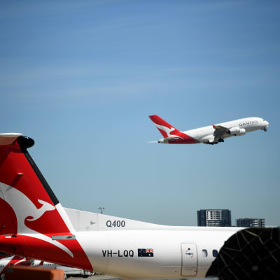 Qantasin lentokoneita lentokentällä. Taustalla lentokone nousemassa ilmaan. 