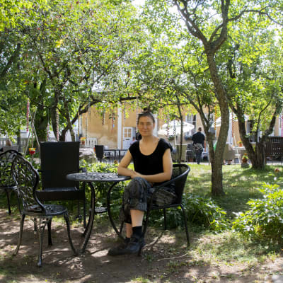 Kvinnan sitter ute vid ett litet bord ute på gården bland träden. Hon har mörka kläder på sig.