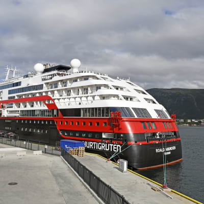Bild på svart, rött, vitt kryssningsfartyg i hamn. I bakgrunden syns berg.