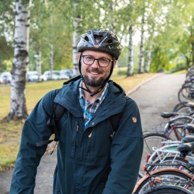 Matti Reinikainen i förd cykelhjälm, rutig skjorta och blå jacka poserar på sin cykel i höstligt landskap.