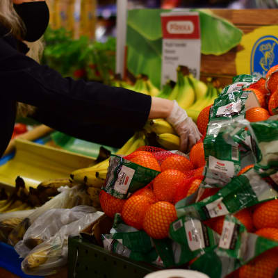 Ett butiksbiträde ordnar bananer och apelsiner med plasthandskar på.