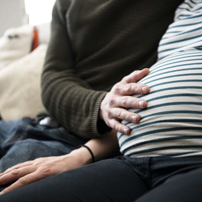 Gravid kvinna sitter på soffa med sin partners hand på magen.