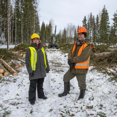 Saija Huuskonen och Markku Rantala står framför en skogsmaskin som håller på att avverka skog.