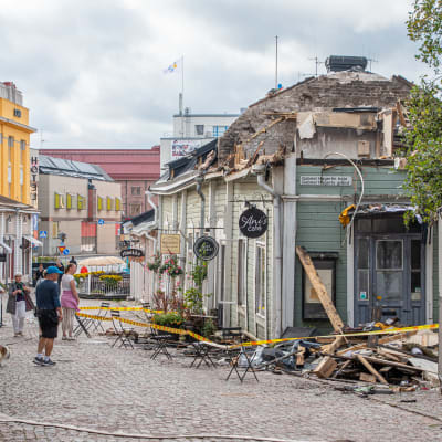 Personer på gata tittar på en träbyggnad som förstörts i en brand. 