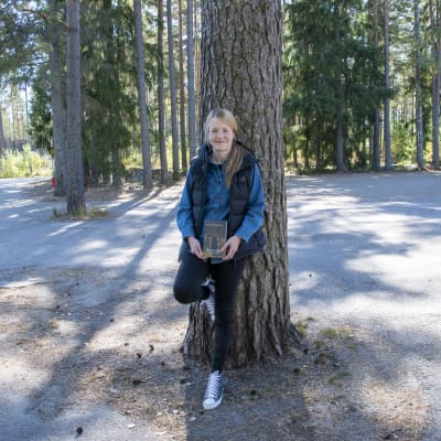 Mirkka Lappalainen lutar mot ett träd och håller i famnen sin bok Smittenin murha.