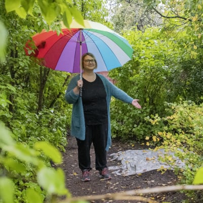 En kvinna med ett färgglatt paraply står i en trädgård och pekar ut ett område med en presenning på.