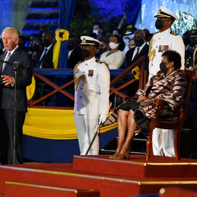 Prins Charles står vid ett podium och håller tal. På en stol på en upphöjning sitter Barbados president Sandra Mason och lyssnar. Omkring dem syns ett antal dignitärer i uniform.