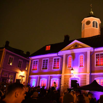En gammal rådhusbyggnad är upplyst med röda lampor. Framför står en stor folkmassa.