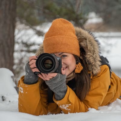 Anette ligger i snön och tar en bild med sin systemkamera. Hon ler. Har en orange jacka och orange mössa. 