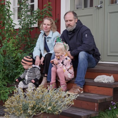 En kvinna och en man sitter på en trappa framför en grön dörr. Framför kvinnan sitter en liten pojke och framför mannen en liten flicka.