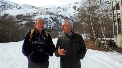 Två gråhåriga män står och diskuterar i ett snöigt landskap.