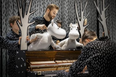 Två dockor föreställande mumintroll kikar fram ovanpå ett piano. 