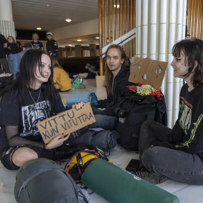 Mielenosoitukseen osallistuvat Humakin opiskelijat Maija Heikkilä ja Noora Hartikainen istuvat yliopiston päärakennuksen aulassa. He ovat varustautuneet tapahtumaan kyltein ja makuupussein.
