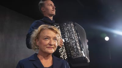En blond kvinna sitter framför en man som spelar dragspel. 