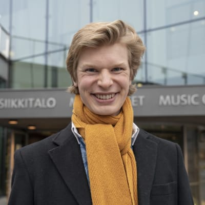 En ung man med stor halsduk utanför ett hus med texten "musiikkitalo".