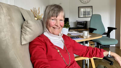 En äldre kvinna i röd tröja sitter i en fåtölj på ett kontor. Hon tittar leende in i kameran.