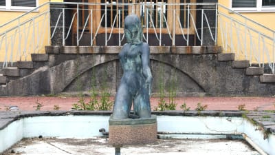 En staty, kvinnlig figur, i en liten ljusblå bassäng utan vatten, sand och löv och vattenrör syns på bassängbottnen.
